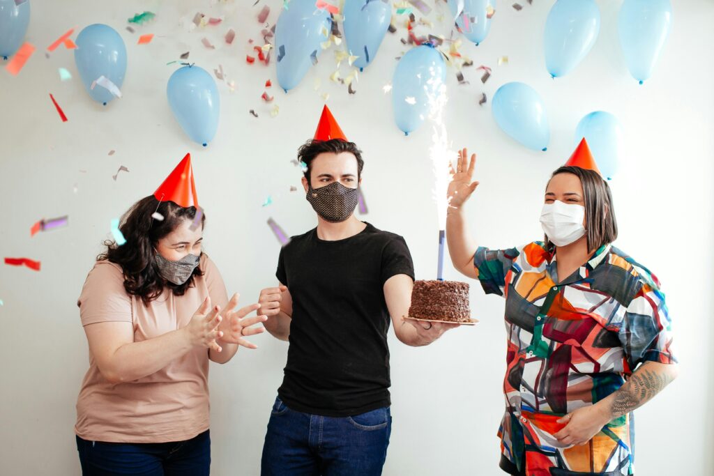Three adults wearing masks celebrate a birthday with cake, balloons, and confetti indoors.